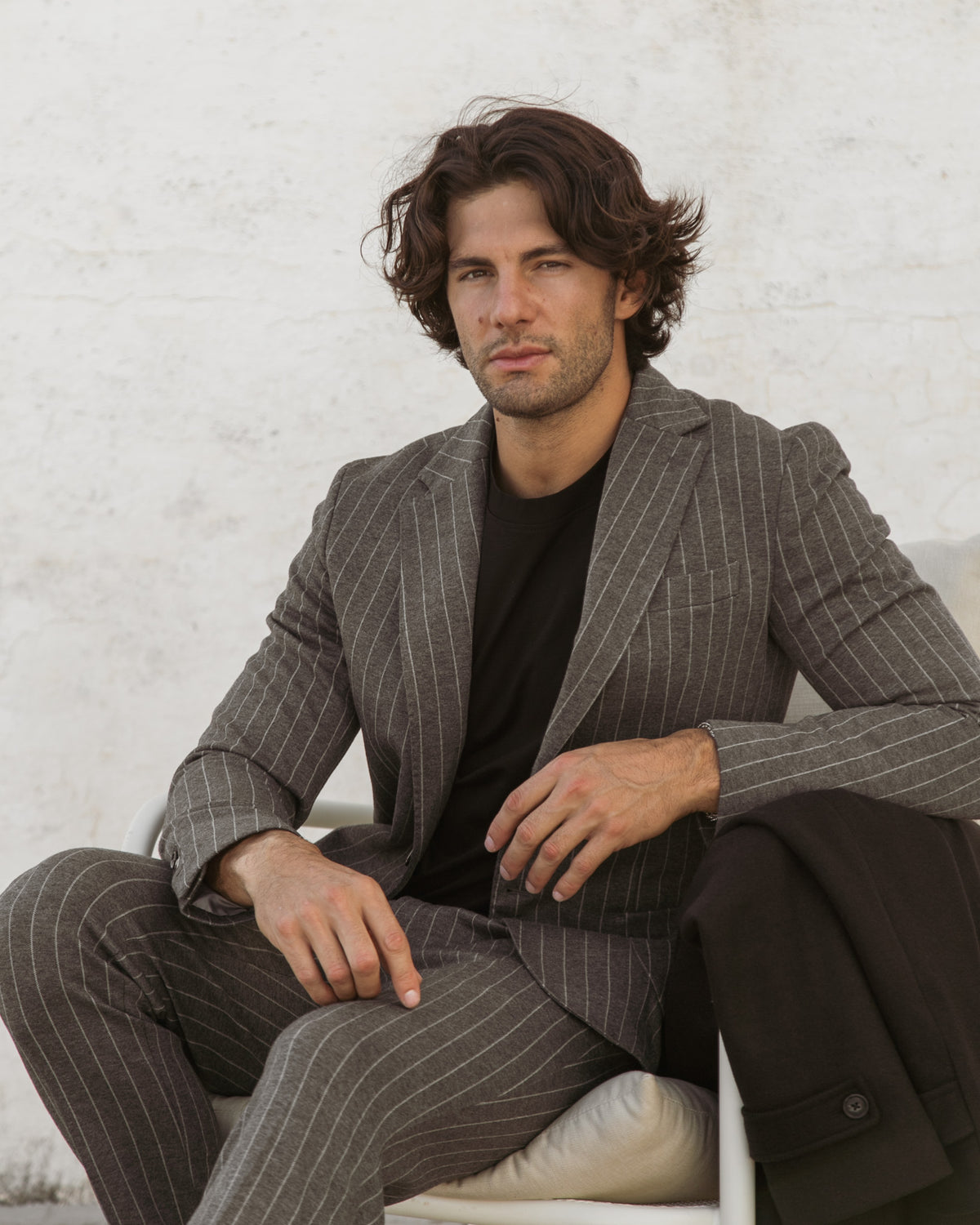 Man in gray pinstripe wool suit with black t-shirt, seated on white chair against white brick wall.