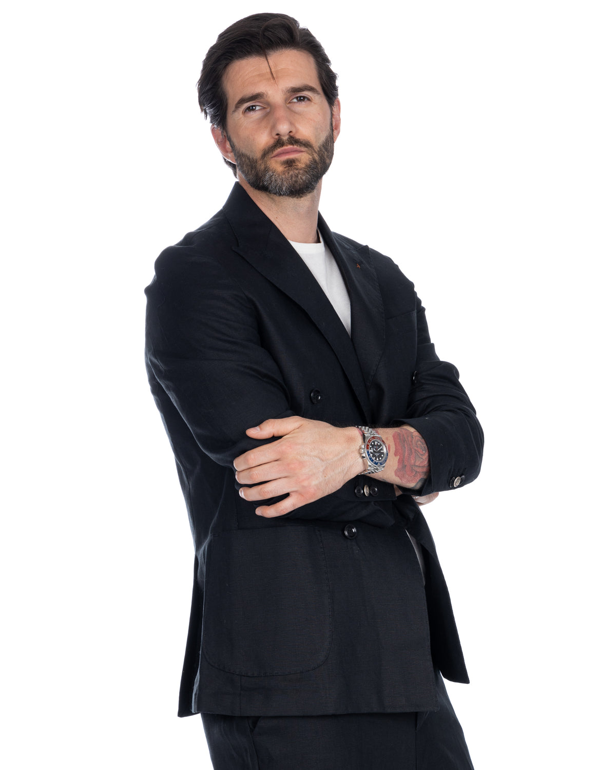 Man wearing black double-breasted wool suit with white t-shirt, silver watch, and arm tattoo, standing with crossed arms.