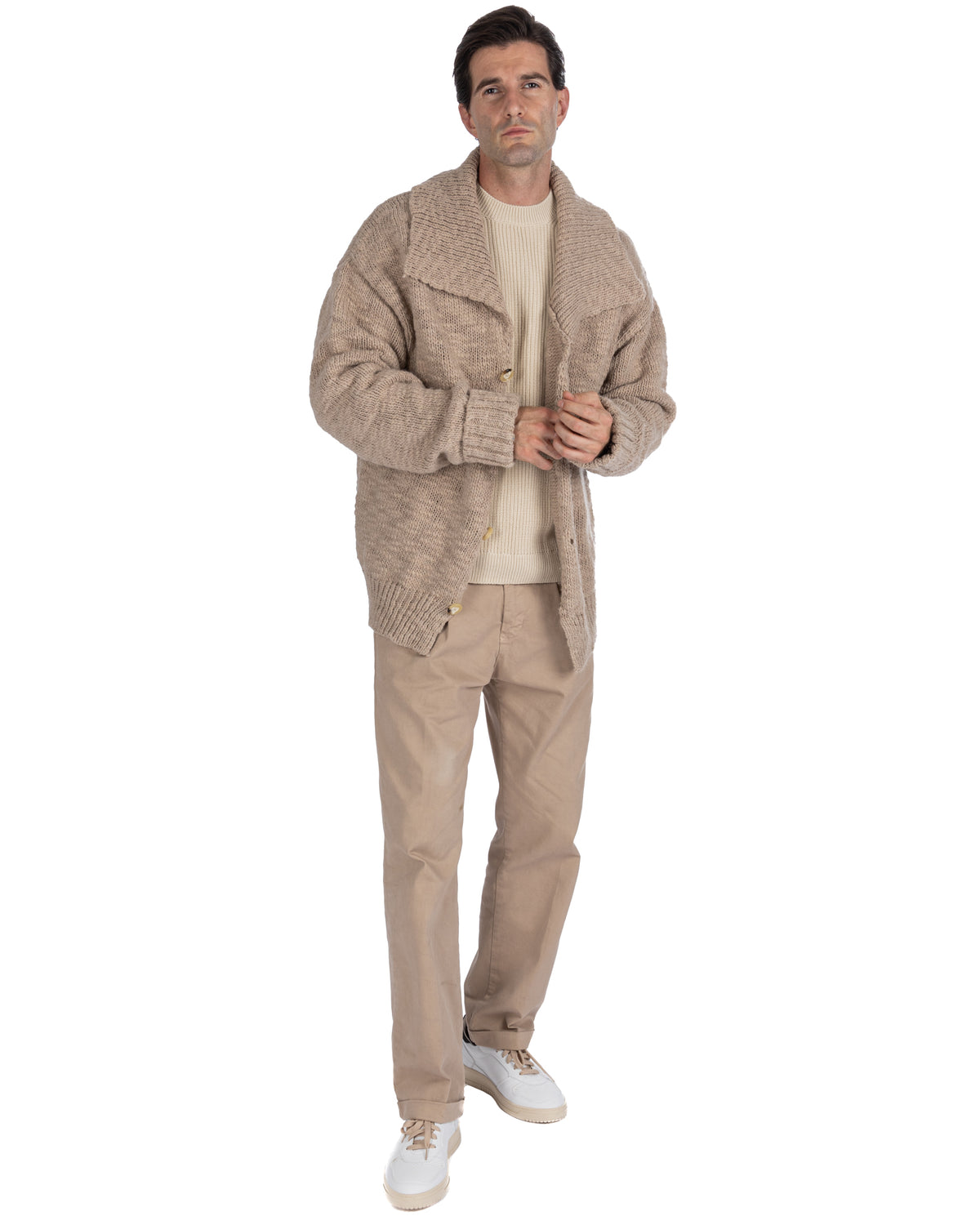 Man wearing beige chunky knit cardigan over cream sweater with tan chinos and white sneakers, full body view.